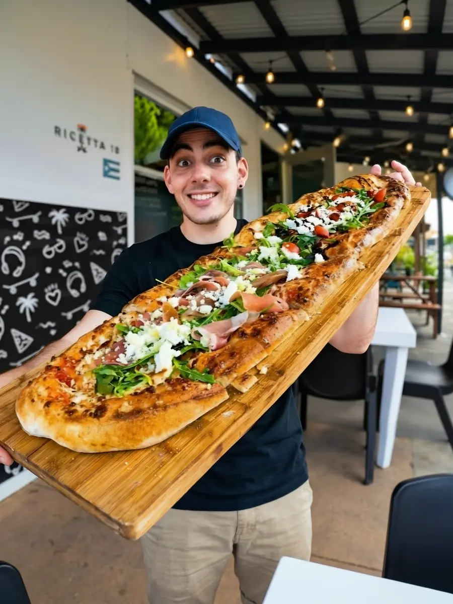 A happy visitor at La Ricetta holding a giant long pizza with prosciutto, arugula, goat cheese and cherry tomatoes